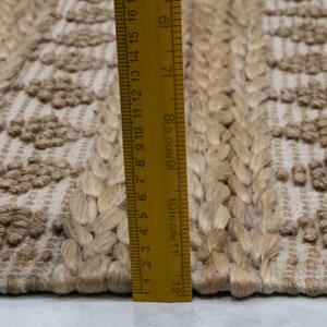 May include: Close-up of a natural fiber rug with a woven pattern. The rug is beige and has a textured surface. A ruler is placed next to the rug, showing the measurement in inches.