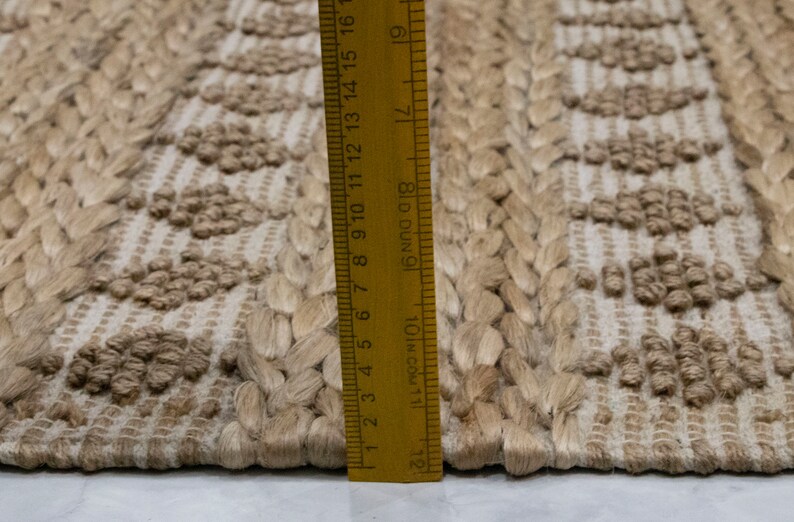 May include: Close-up of a natural fiber rug with a woven pattern. The rug is beige and has a textured surface. A ruler is placed next to the rug, showing the measurement in inches.