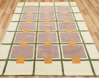 Op de afbeelding: Rechthoekig vloerkleed met een crèmekleurige achtergrond en een rasterpatroon. Het raster wordt gevormd door groene lijnen, mauve vierkanten en oranje accenten. Het vloerkleed ligt op een houten vloer.