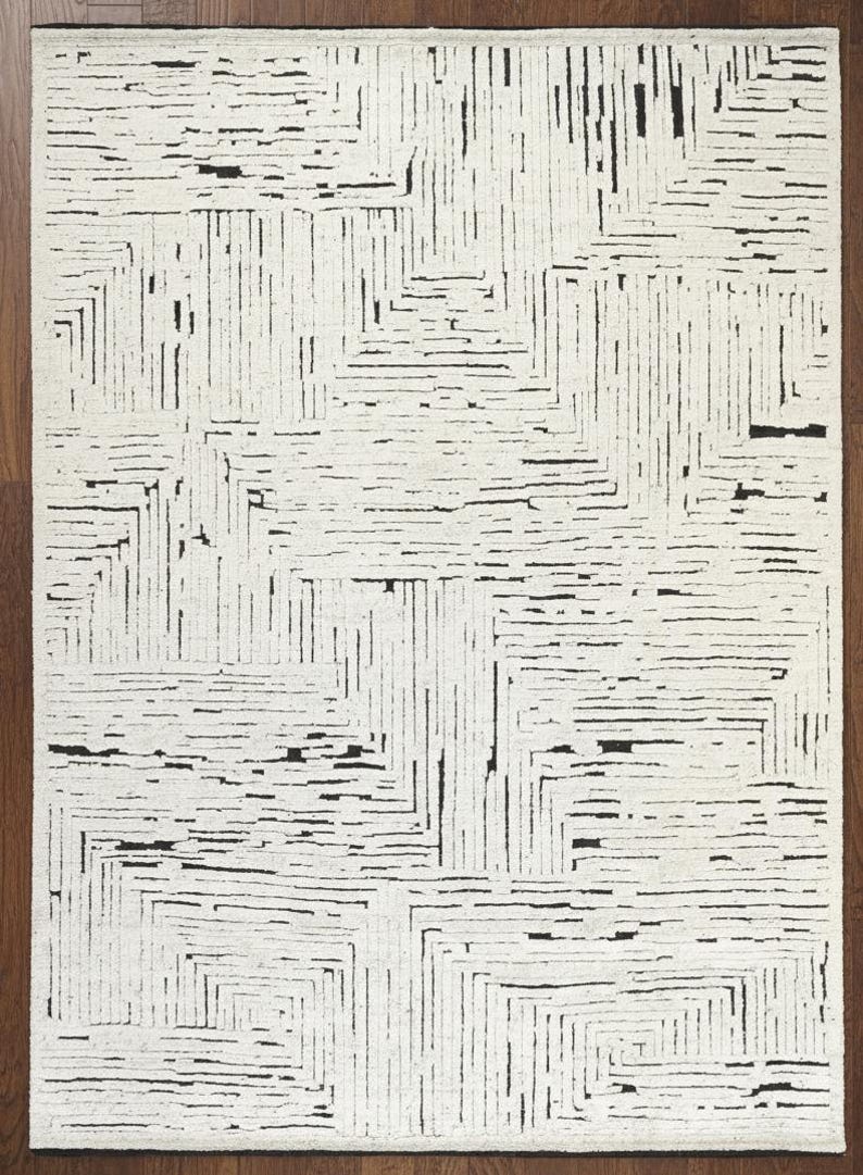 May include: Rectangular area rug with a geometric pattern. The rug features a white background with a repeating design of black lines and rectangular shapes. The rug is made of a soft material and is suitable for living rooms and bedrooms.