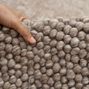 May include: Close-up of a beige and brown wool rug with a textured, bubble-like surface. The rug is made of small, round balls of wool that are tightly woven together.