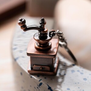 May include: A small, antique-style coffee grinder keychain made of metal with a copper finish. The grinder has a black handle and a silver chain.