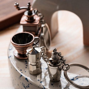 May include: A set of five silver and copper-toned miniature coffee-themed keychains. The keychains include a coffee grinder, a coffee filter, a moka pot, a shaker, and a ring.