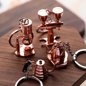 Könnte beinhalten: Vier roségoldene Kaffeetassen-Anhänger aus Metall. Die Anhänger sind eine Kaffeemühle, eine Kaffeekanne, eine French Press und ein Wasserkocher. Die Anhänger befinden sich auf einer Holzoberfläche.