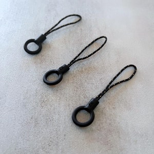 Può includere: Tre portachiavi in ​​cordino nero e oro con anelli in plastica nera. I portachiavi sono realizzati con un cordino sottile e intrecciato.