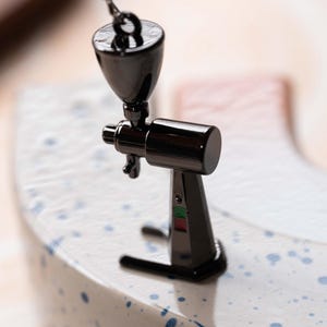May include: A miniature black metal coffee grinder with a silver chain hanging from the top. The grinder is sitting on a white surface with blue speckles.