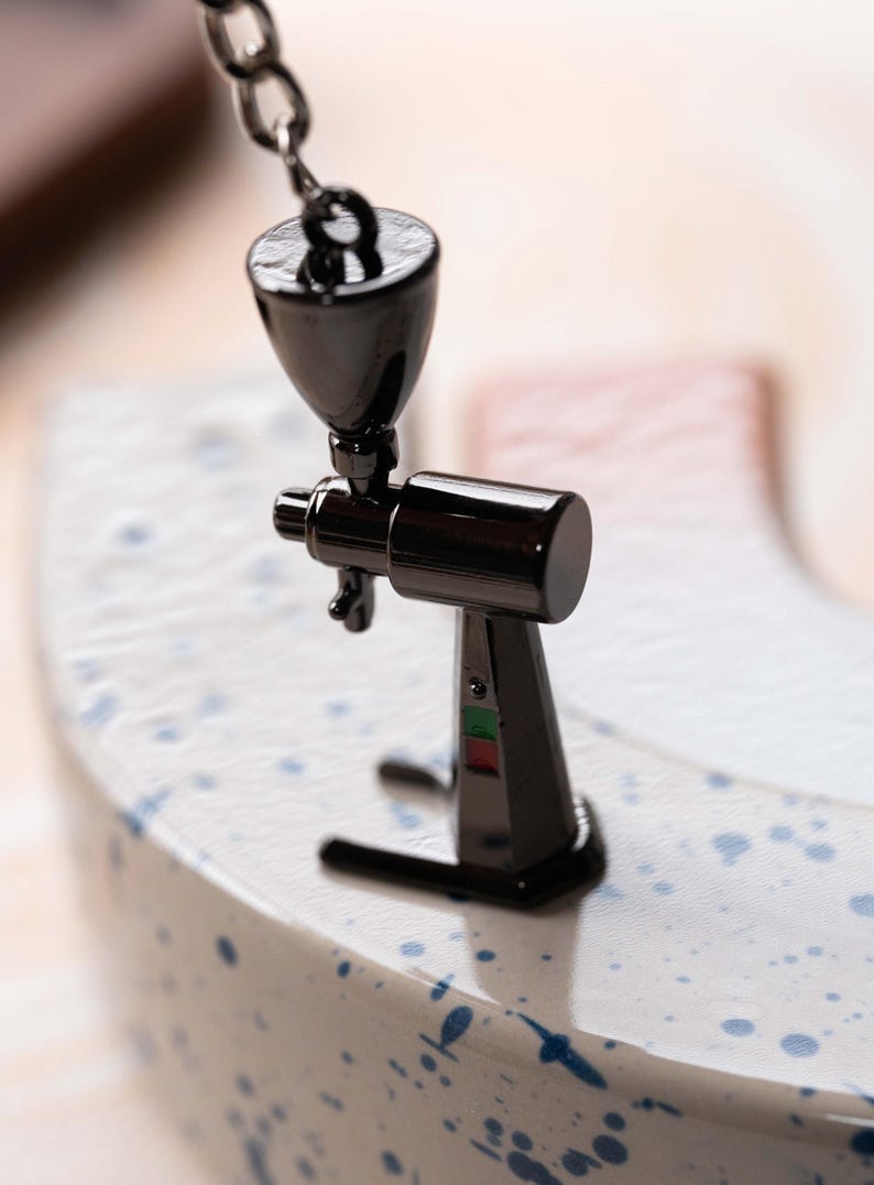 May include: A miniature black metal coffee grinder with a silver chain hanging from the top. The grinder is sitting on a white surface with blue speckles.