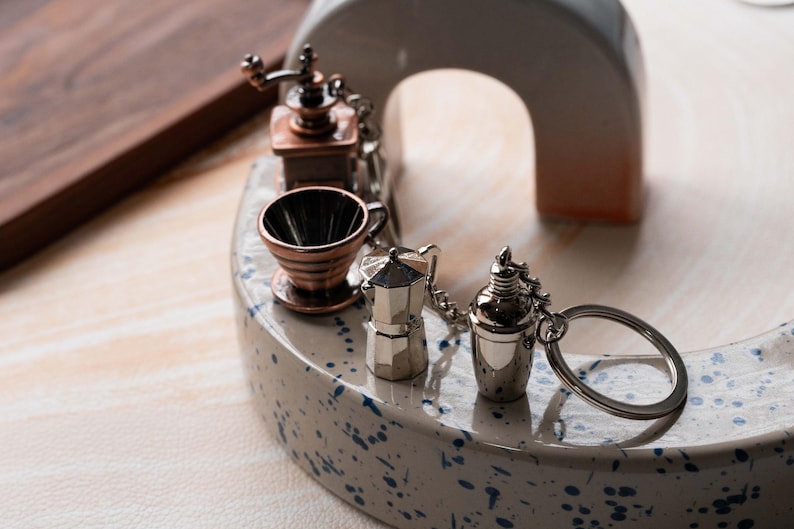 May include: A set of five miniature coffee-themed keychains, including a grinder, a filter, a coffee pot, a shaker, and a ring. The keychains are silver and copper in color and are displayed on a white ceramic surface with blue speckles.