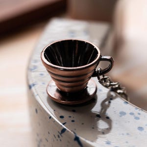 May include: A small, copper-colored coffee filter with a handle and a chain. The filter is sitting on a white surface with blue speckles.