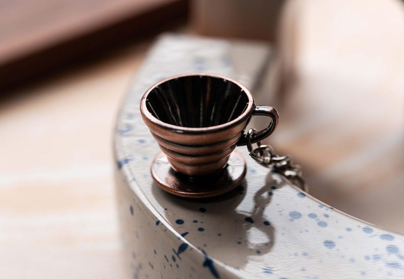 May include: A small, copper-colored coffee filter with a handle and a chain. The filter is sitting on a white surface with blue speckles.