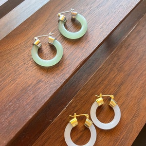 May include: Four pairs of hoop earrings. Two pairs are a light green color with silver-colored clasps. The other two pairs are white with gold-colored clasps. The earrings are displayed on a dark brown wooden surface.
