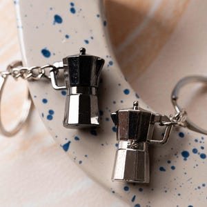 May include: Two silver metal keychains shaped like coffee pots. The keychains are attached to a silver metal ring. The keychains are on a white surface with blue splatter paint.