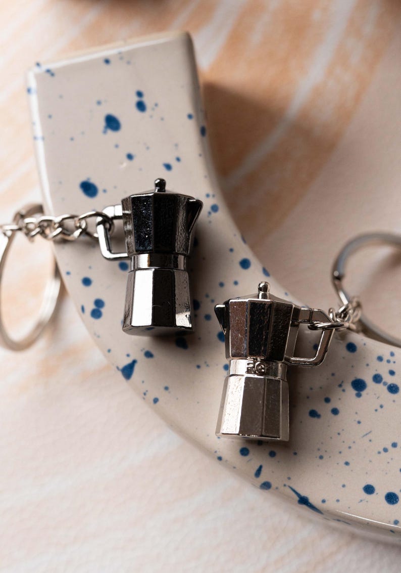 May include: Two silver metal keychains shaped like coffee pots. The keychains are attached to a silver metal ring. The keychains are on a white surface with blue splatter paint.