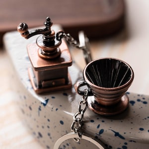 May include: A silver keychain with a miniature coffee grinder and a coffee filter charm. The grinder is made of copper and has a black handle. The filter is also made of copper and has a black interior.