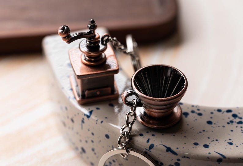 May include: A silver keychain with a miniature coffee grinder and a coffee filter charm. The grinder is made of copper and has a black handle. The filter is also made of copper and has a black interior.
