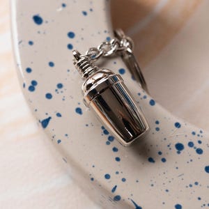 May include: A silver metal keychain shaped like a cocktail shaker. The keychain is on a white surface with blue splatter paint.