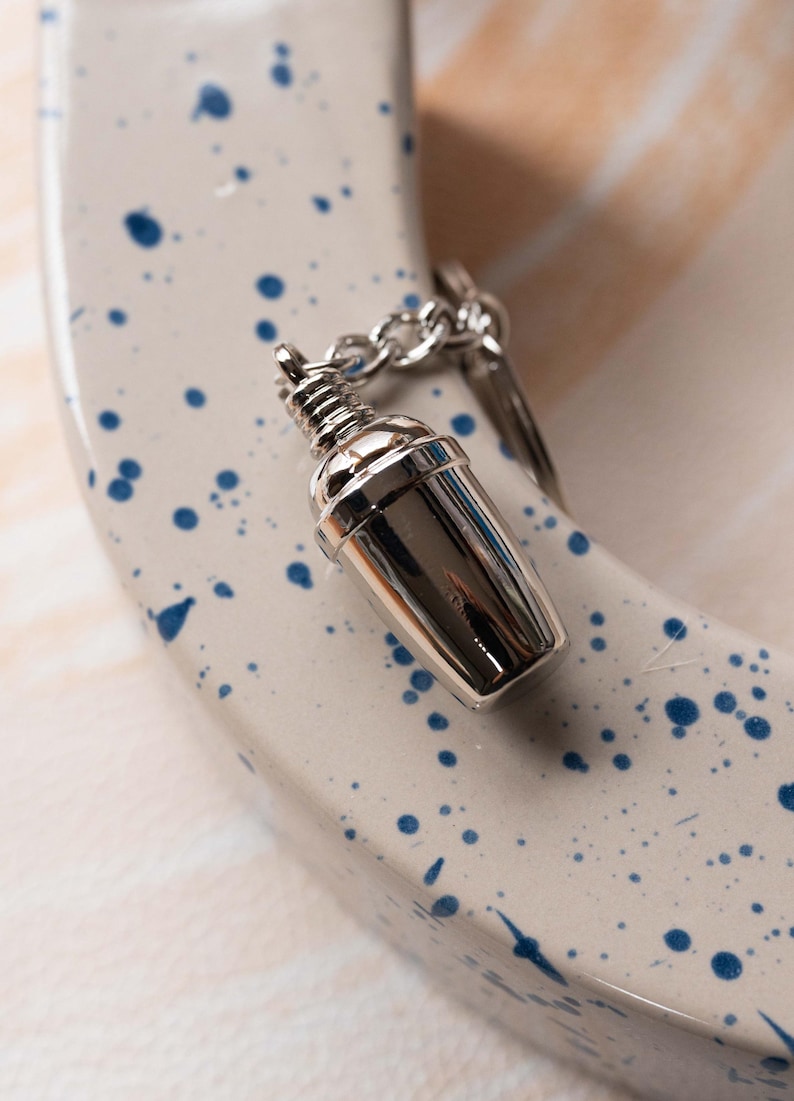 May include: A silver metal keychain shaped like a cocktail shaker. The keychain is on a white surface with blue splatter paint.