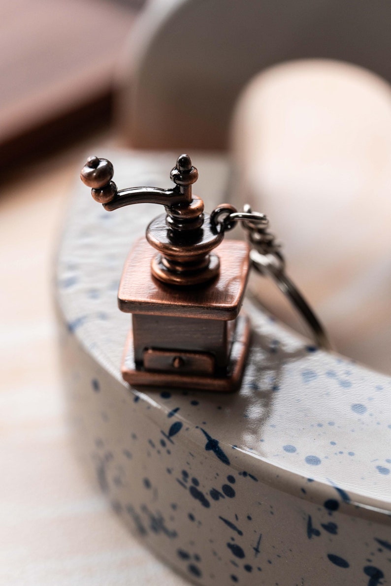 May include: A small, antique-style coffee grinder keychain made of metal with a copper finish. The grinder has a black handle and a silver chain.