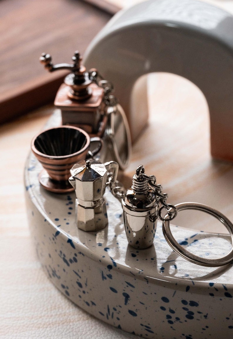 May include: A set of five silver and copper-toned miniature coffee-themed keychains. The keychains include a coffee grinder, a coffee filter, a moka pot, a shaker, and a ring.