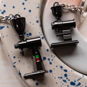 May include: Two black metal coffee grinder keychain charms. One charm has a green and red accent. The charms are on a white and blue speckled surface.