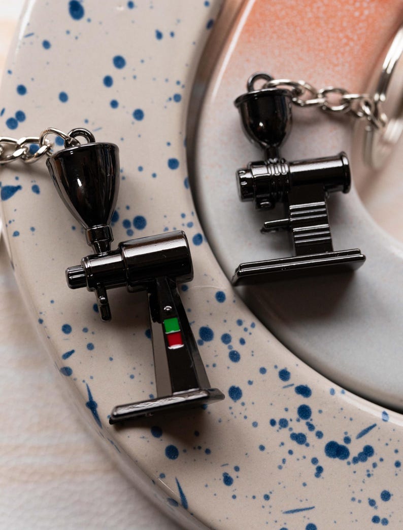 May include: Two black metal coffee grinder keychain charms. One charm has a green and red accent. The charms are on a white and blue speckled surface.