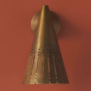 May include: A brass wall sconce with a conical shade. The shade has vertical and circular cutouts. The sconce is mounted on a circular base against a reddish-orange wall. The light fixture has a vintage design.