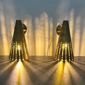 May include: Two brass cone-shaped wall sconces with a series of vertical and circular cutouts. The sconces are lit from within, casting a warm glow and creating a pattern of light and shadow on the wall. The sconces are mounted on round brass backplates.
