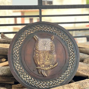 May include: A round, wooden wall hanging featuring a detailed owl carving. The owl is depicted in a central medallion, with intricate feather and branch details. The outer ring has a woven design, and the color palette includes brown and gold tones.