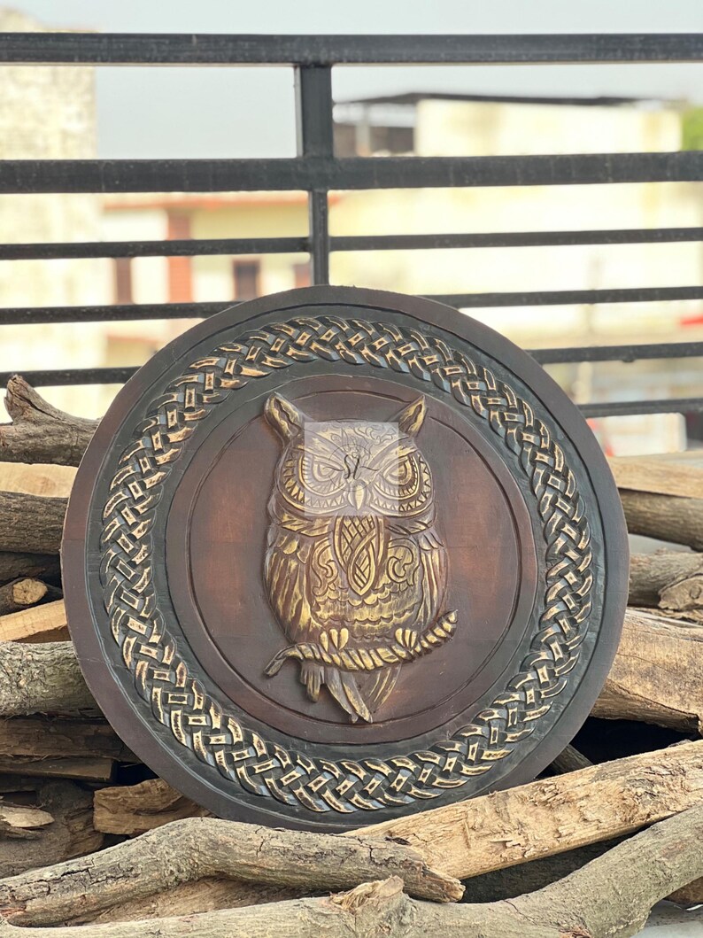 May include: A round, wooden wall hanging featuring a detailed owl carving. The owl is depicted in a central medallion, with intricate feather and branch details. The outer ring has a woven design, and the color palette includes brown and gold tones.