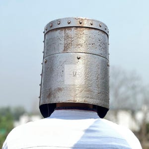 K&ouml;nnte beinhalten: Ein silberfarbener Metallhelm, vermutlich ein Ritterhelm, mit zylindrischer Form und genieteter Konstruktion. Der Helm hat ein verwittertes Aussehen. Das Bild ist aus einem niedrigen Winkel aufgenommen und zeigt den R&uuml;cken der Person, die den Helm tr&auml;gt.