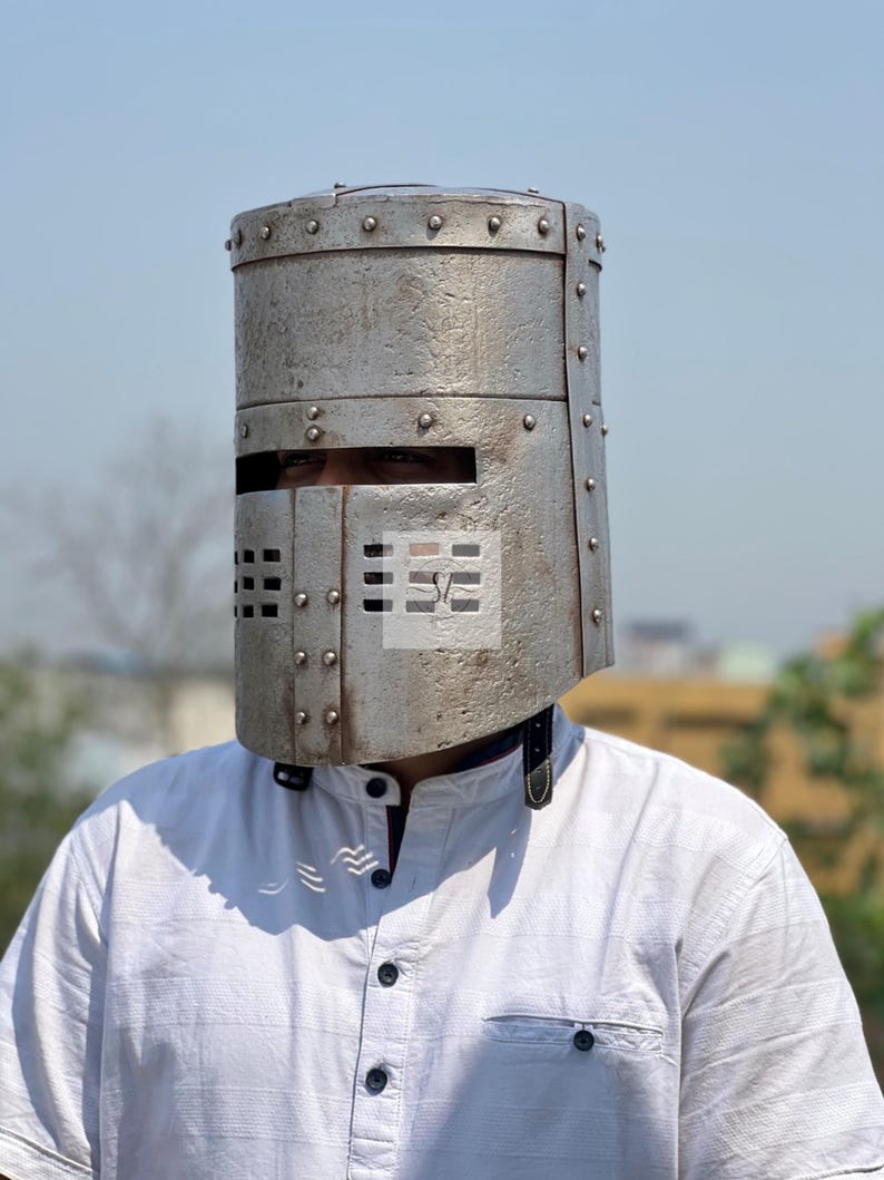 K&ouml;nnte beinhalten: Ein silberfarbener mittelalterlicher Ritterhelm mit zylindrischer Form und genieteter Konstruktion. Der Helm hat einen horizontalen Augenschlitz und rechteckige Bel&uuml;ftungsl&ouml;cher. Die Person tr&auml;gt ein wei&szlig;es Hemd mit Kn&ouml;pfen.
