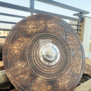 May include: A round, wooden shield with intricate carvings and a central metal boss. The shield features detailed patterns and designs, with a dark brown finish. The shield is displayed against a backdrop of wood and a metal railing.