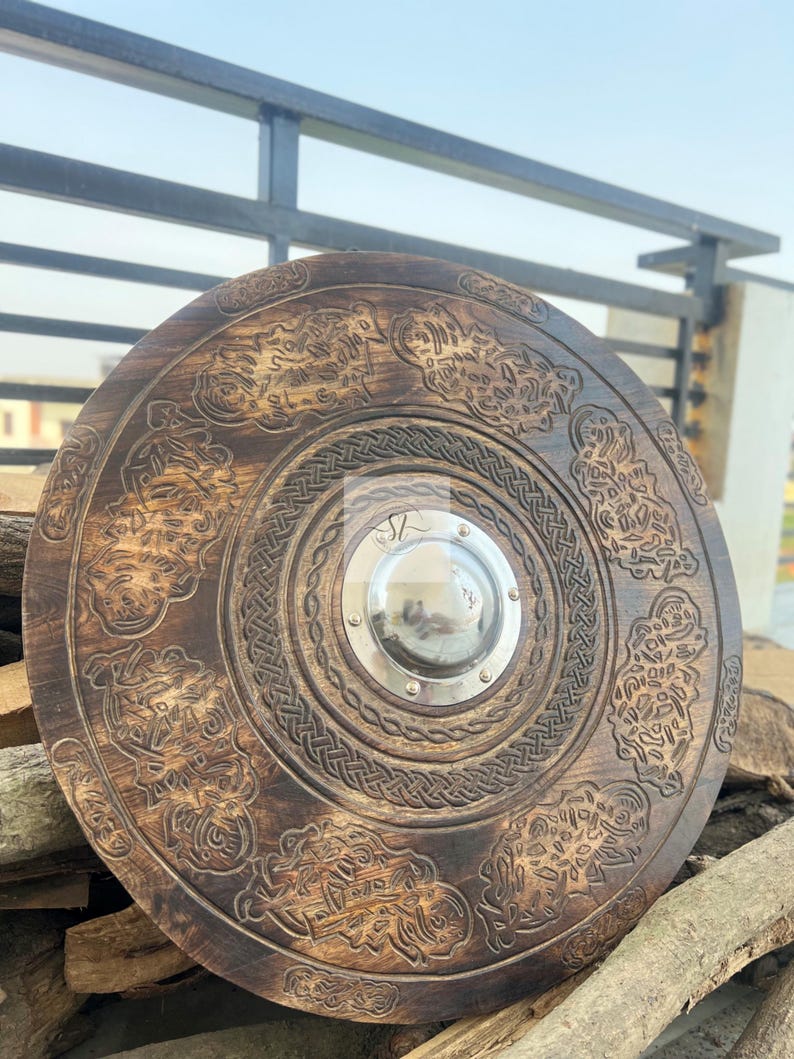 May include: A round, wooden shield with intricate carvings and a central metal boss. The shield features detailed patterns and designs, with a dark brown finish. The shield is displayed against a backdrop of wood and a metal railing.
