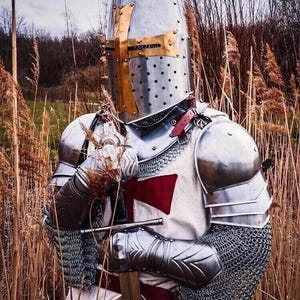 Puede incluir: Un caballero con armadura completa, con un casco con una cruz dorada, se encuentra en un campo de hierba alta. El caballero sostiene una espada en su mano derecha y tiene una cruz roja en su pecho.