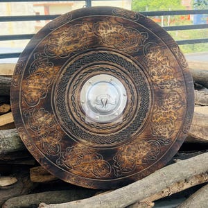 May include: A large, round, wooden shield with intricate carved designs and a central metal plate. The shield is dark brown with detailed patterns around the edges and center. The metal plate has the initials "SL".