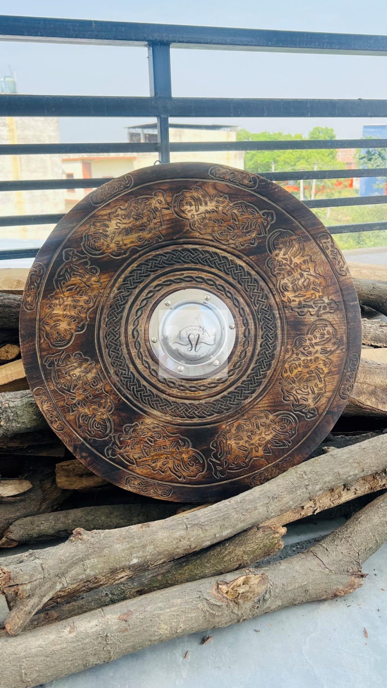 May include: A large, round, wooden shield with intricate carved designs and a central metal plate. The shield is dark brown with detailed patterns around the edges and center. The metal plate has the initials "SL".