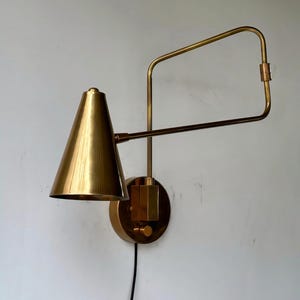 Puede incluir: Aplique de pared de latón con una pantalla cónica y un brazo rectangular. El aplique está montado sobre una base circular con un pequeño interruptor. Un cable de alimentación negro sale de la base. La lámpara es de color dorado cálido.