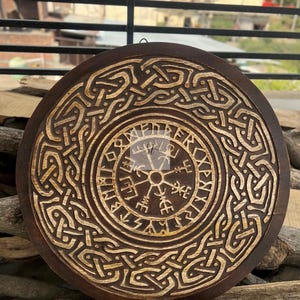 May include: Round, wooden wall art featuring a detailed, carved Viking design. The dark brown wood contrasts with the lighter, carved patterns, including a central compass symbol and intricate knotwork. A unique, decorative piece.