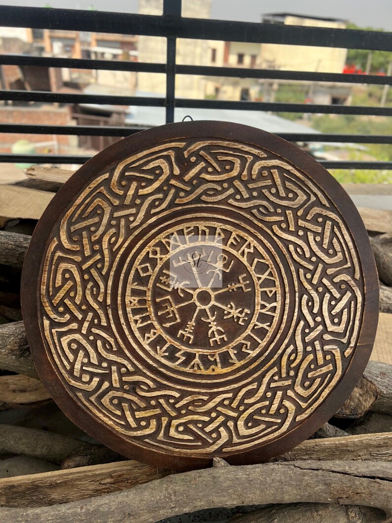May include: Round, wooden wall art featuring a detailed, carved Viking design. The dark brown wood contrasts with the lighter, carved patterns, including a central compass symbol and intricate knotwork. A unique, decorative piece.