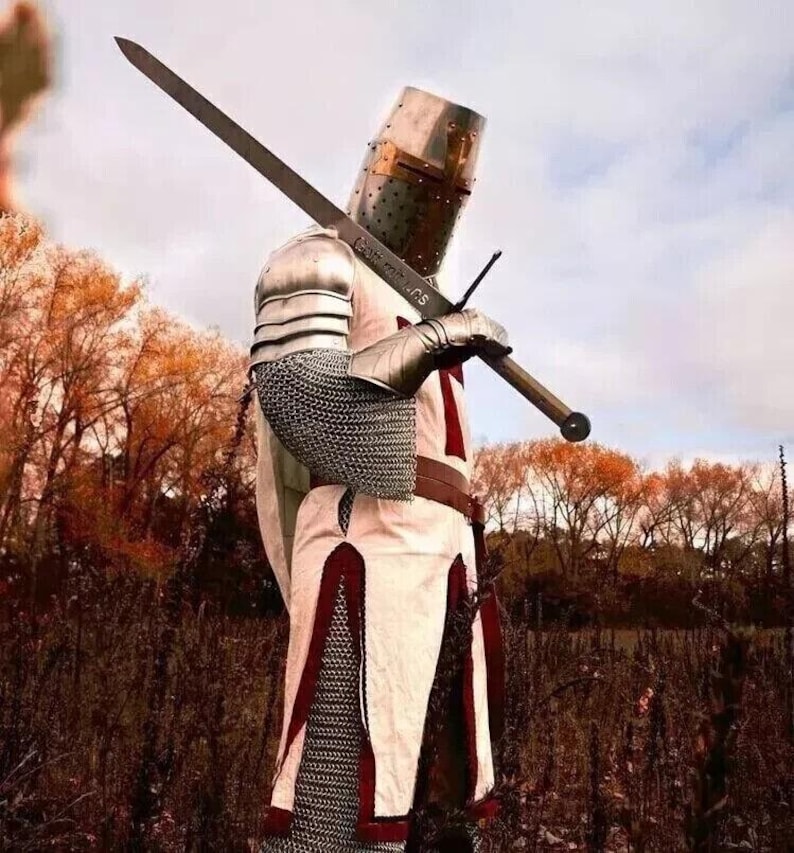 Puede incluir: Un caballero con armadura completa, vistiendo una t&uacute;nica blanca con una cruz roja, sostiene una espada a trav&eacute;s de su cuerpo. Est&aacute; de pie en un campo con &aacute;rboles en el fondo.