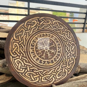 May include: A round, wooden wall hanging with a carved Viking compass design. The dark brown wood features intricate knotwork and a lighter, natural wood finish in the carved details. The piece is approximately 12 inches in diameter.