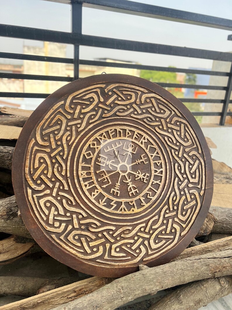 May include: A round, wooden wall hanging with a carved Viking compass design. The dark brown wood features intricate knotwork and a lighter, natural wood finish in the carved details. The piece is approximately 12 inches in diameter.