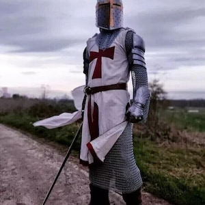 Puede incluir: Un caballero con armadura completa, vistiendo una t&uacute;nica blanca con una cruz roja, se encuentra en un camino de tierra. Lleva una espada y tiene un pa&ntilde;o blanco drapeado sobre su hombro.