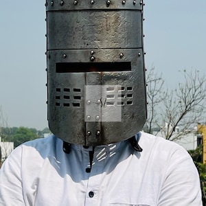 K&ouml;nnte beinhalten: Ein verwitterter, genieteter Ritterhelm aus Metall mit rechteckigem Visier und Bel&uuml;ftungsl&ouml;chern. Der Helm ist zylindrisch. Die Person, die ihn tr&auml;gt, tr&auml;gt ein wei&szlig;es Hemd. Der Hintergrund ist ein klarer blauer Himmel.