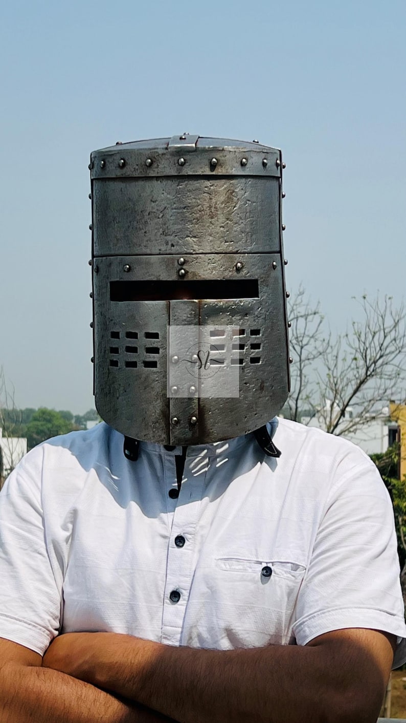 K&ouml;nnte beinhalten: Ein verwitterter, genieteter Ritterhelm aus Metall mit rechteckigem Visier und Bel&uuml;ftungsl&ouml;chern. Der Helm ist zylindrisch. Die Person, die ihn tr&auml;gt, tr&auml;gt ein wei&szlig;es Hemd. Der Hintergrund ist ein klarer blauer Himmel.