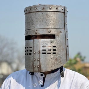 K&ouml;nnte beinhalten: Ein silberfarbener Metallhelm mit zylindrischem Oberteil und rechteckigen Augen- und Mund&ouml;ffnungen. Der Helm ist genietet und wird &uuml;ber einem wei&szlig;en Hemd getragen. Es handelt sich wahrscheinlich um ein historisches oder Kost&uuml;mteil.