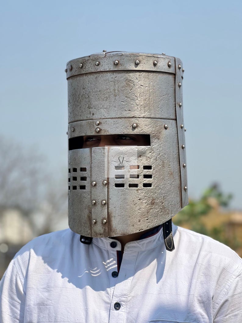 K&ouml;nnte beinhalten: Ein silberfarbener Metallhelm mit zylindrischem Oberteil und rechteckigen Augen- und Mund&ouml;ffnungen. Der Helm ist genietet und wird &uuml;ber einem wei&szlig;en Hemd getragen. Es handelt sich wahrscheinlich um ein historisches oder Kost&uuml;mteil.