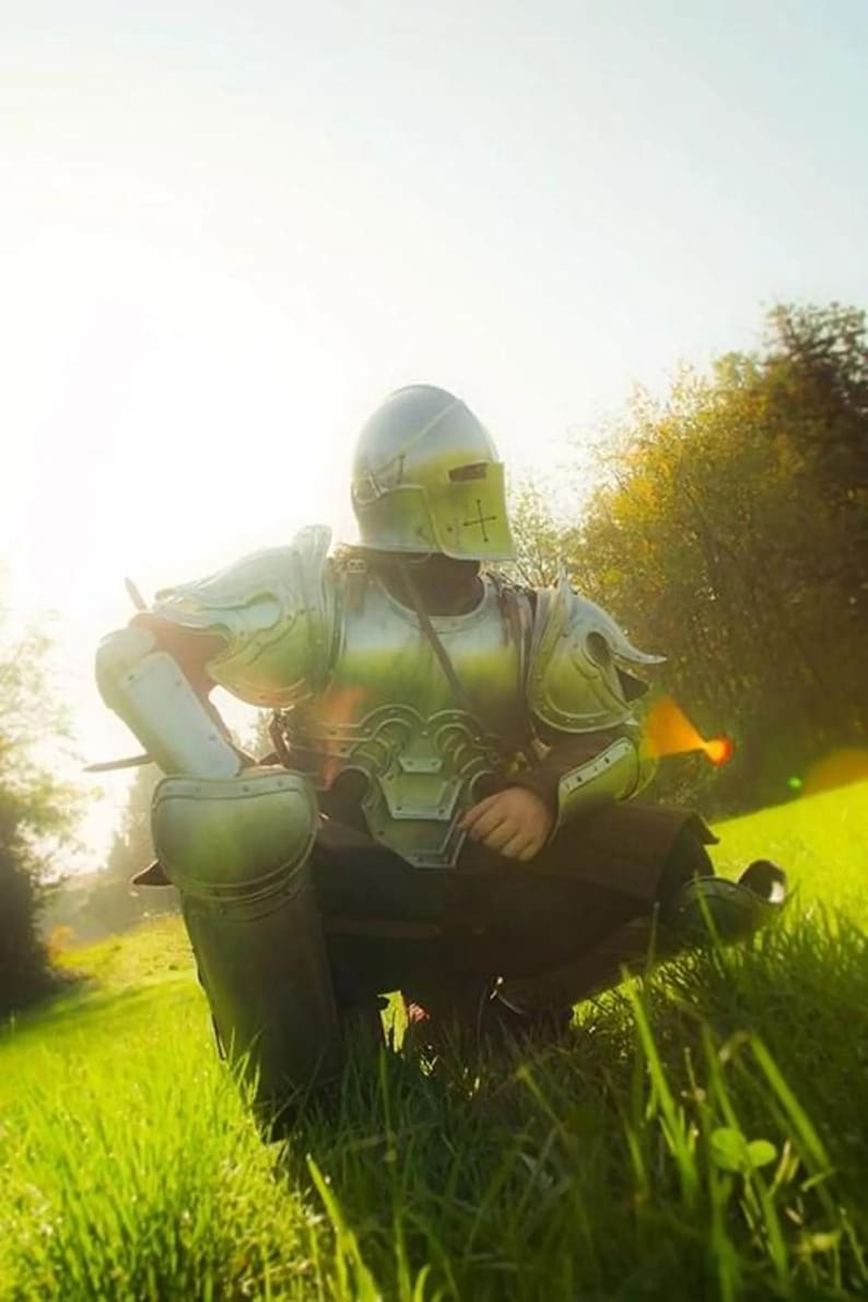 K&ouml;nnte beinhalten: Eine Person in silberfarbener mittelalterlicher R&uuml;stung, einschlie&szlig;lich eines Helms mit Kreuz, sitzt in einem grasbewachsenen Feld. Die R&uuml;stung reflektiert das Sonnenlicht und erzeugt eine historische und fantastische Szene.
