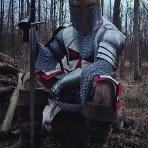 Puede incluir: Un caballero con armadura completa, vistiendo una t&uacute;nica de malla y un casco con una cruz roja, est&aacute; sentado en un entorno boscoso. Sostiene una espada en su mano derecha.