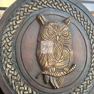 May include: A round, wooden wall hanging featuring a detailed owl carving. The owl is perched on a branch, with intricate feather and facial details. The design is framed by a braided border, with a dark brown and gold color scheme.